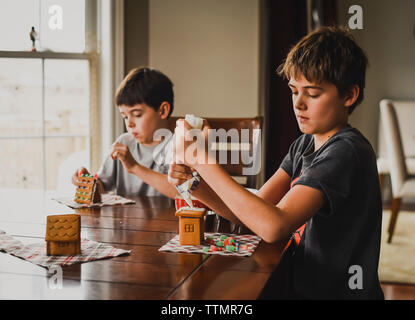 Zwei Jungen dekorieren Lebkuchenhäuser zusammen am Tisch. Stockfoto