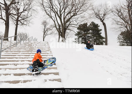 Zwei Jungen rodeln auf einem verschneiten Hügel in einem Park an einem Wintertag. Stockfoto