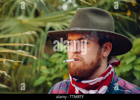 Nahaufnahme des bärtigen jungen Mann mit Hut und Halstuch beim Rauchen Zigarette Stockfoto