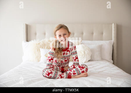 Lächelnde blonde lockige blonde Mädchen mit Urlaub Schlafanzug auf weißen Bett Stockfoto