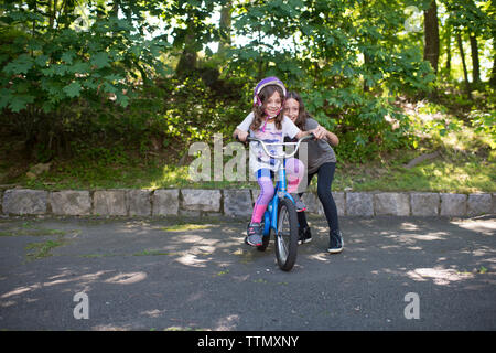 Mädchen Unterstützung Schwester im Reiten Fahrrad auf der Straße gegen Bäume im Park Stockfoto