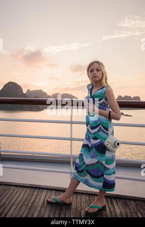 Portrait von nettes Mädchen mit Kamera stehend durch Geländer auf Boot im Meer gegen Himmel bei Sonnenuntergang Stockfoto