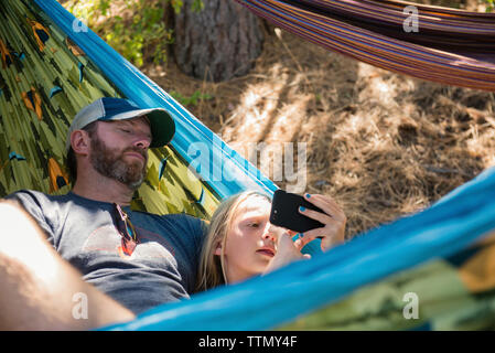 Hohe Betrachtungswinkel der Tochter mit Handy beim Lügen mit Vater in der Hängematte am Wald Stockfoto