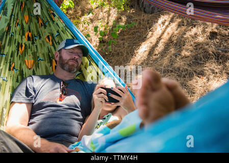 Hohe Betrachtungswinkel der Tochter mit Smart Phone beim Lügen mit Vater in der Hängematte am Wald Stockfoto