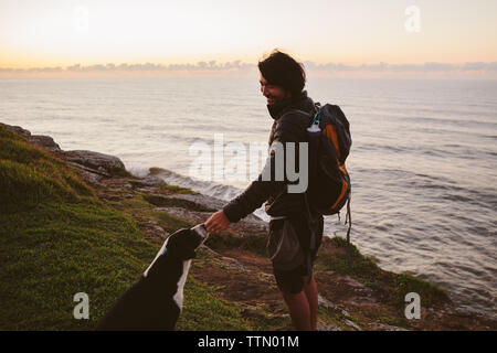 Seitenansicht der männlichen Wanderer berühren Hund beim Stehen auf einem Hügel mit Meerblick Stockfoto
