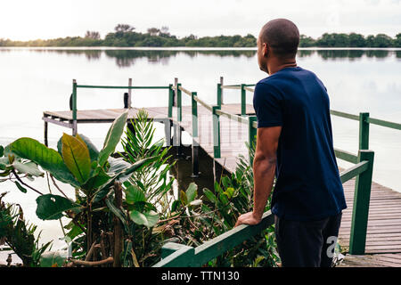 Junger Mann auf der Suche am See beim Stehen auf Pier von Pflanzen bei Sonnenuntergang Stockfoto