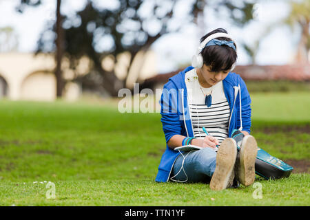 Volle Länge des Jugendmädchen schreiben auf Journal während der Wiedergabe von Musik im Park Stockfoto