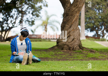 Junges Mädchen schreiben auf Journal während der Wiedergabe von Musik im Park Stockfoto