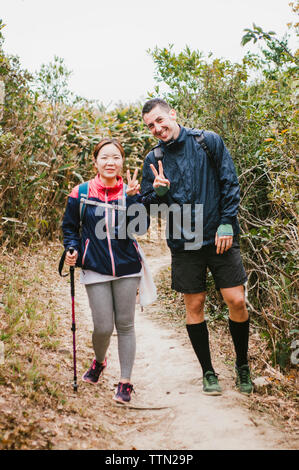 Portrait von glücklich, Freunde, Frieden Zeichen beim Stehen auf Wanderweg inmitten von Pflanzen gegen Sky Stockfoto