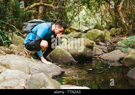 Männliche Wanderer Trinkwasser aus See während kauert auf Felsen im Wald Stockfoto