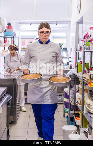 Portrait der männlichen Bäcker Kuchen halten, während die weiblichen Mitarbeiter arbeiten im Hintergrund bei Labor Stockfoto
