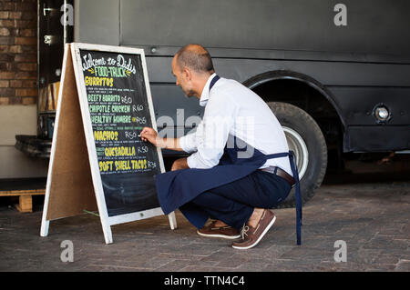 Volle Länge des männlichen Anbieter schreiben auf Tafel während geduckt auf der Straße, die durch Nahrung Lkw Stockfoto