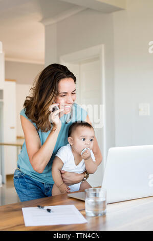 Mutter am Telefon sprechen, beim Tragen von baby boy auf Laptop Stockfoto