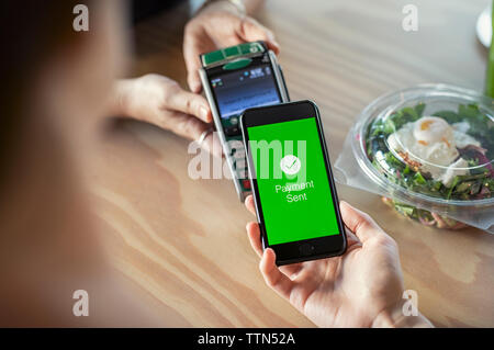 7/8 Hände der weiblichen Kunden mobile Payment an Eigentümer auf hölzernen Tisch in der Cafeteria Stockfoto