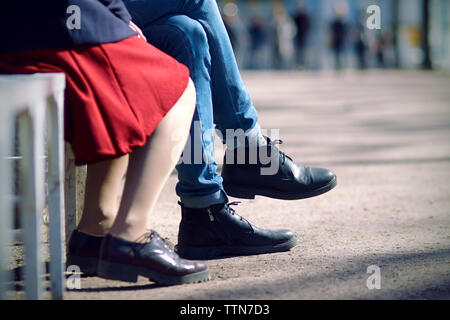 Beine von einem jungen Paar, sitzen an einem Sommertag auf einer Bank. Der Mann ist in engen Jeans und schwarze Stiefel gekleidet, und die Frau im roten Rock und Blackcamo Stockfoto