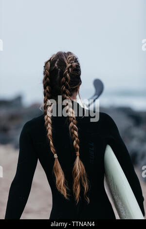 Ansicht der Rückseite Frau mit geflochtenem Haar lang Surfboard Durchführung beim Spaziergang am Strand gegen Sky Stockfoto