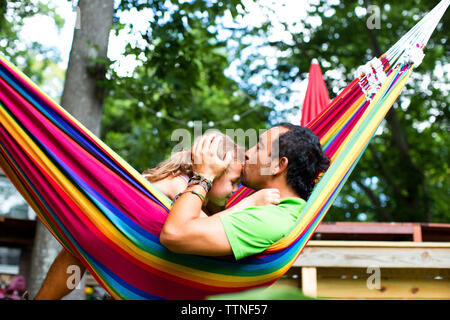 Mann küssen Tochter auf die Stirn, während Sie sich in der Hängematte im Garten Stockfoto