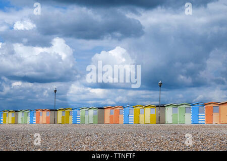 Eine Linie von 22 bunten Badekabinen, die durch die Mitte des Bildes, ist unten ein gelbes Kieselstrand und oben ist ein dramatischer blaue und weiße Bewölkt Stockfoto