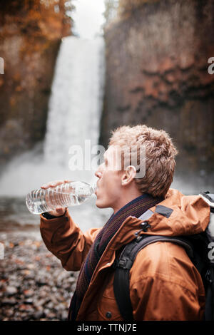 Seitenansicht der männlichen Wanderer Trinkwasser aus der Flasche beim Stehen gegen Wasserfall Stockfoto