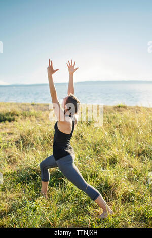 Frau üben warrior Pose auf der Wiese durch das Meer gegen den klaren Himmel Stockfoto