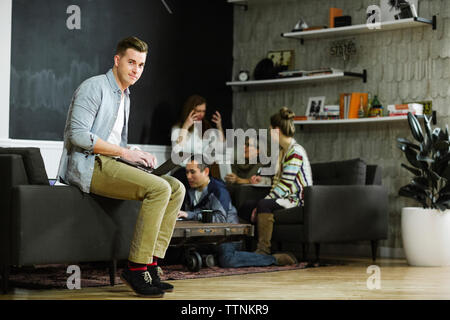 Portrait von Geschäftsmann mit Laptop Computer, während Kollegen sprechen im Hintergrund im Büro Stockfoto