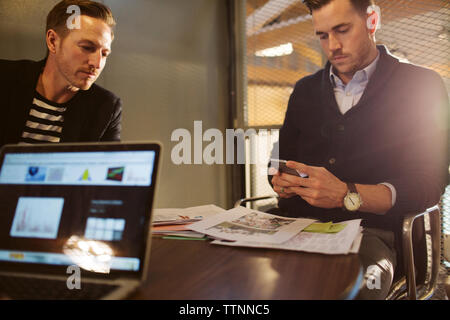 Geschäftsmann benutzt Handy, während er mit männlichem Kollegen im Büro sitzt Stockfoto