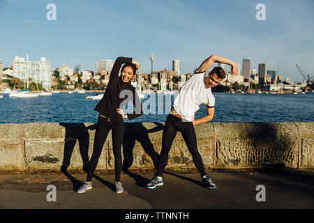 Volle Länge des glückliches junges Paar stretching während des Trainings an der Promenade gegen Stadtbild Stockfoto