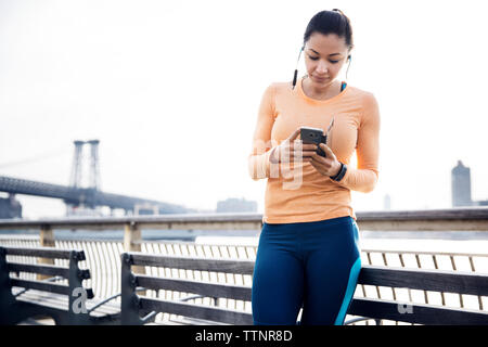 Sportliche Frau Musik hören durch Smart Phone mit Williamsburg Bridge im Hintergrund Stockfoto