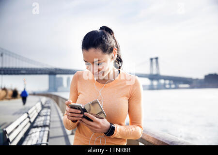 Glückliche athletin Musik hören durch Smart Phone mit Williamsburg Bridge im Hintergrund Stockfoto