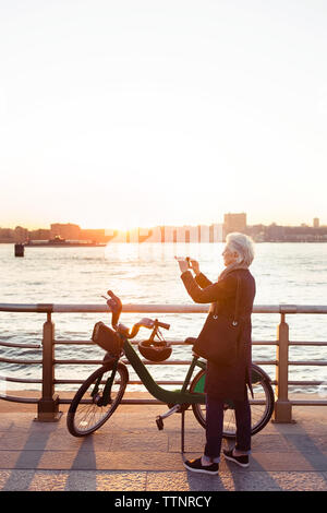 Die volle Länge der älteren Frau fotografieren beim Stehen auf der Promenade bei Sonnenuntergang Stockfoto