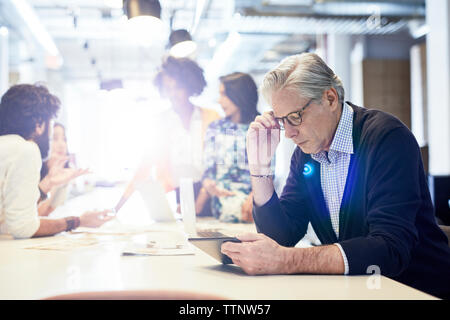 Senior Geschäftsmann mit Tablet Computer mit Geschäftsleuten in Hintergrund arbeiten Stockfoto