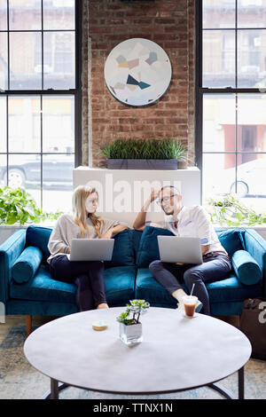 Geschäftskollegen mit Laptops diskutieren, während sie auf dem Sofa vor den Fenstern sitzen Stockfoto