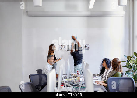 Geschäftsleute diskutieren am Konferenztisch, während Kollegen im Büro Fotoausdrucke an der Pinnwand anbringen Stockfoto