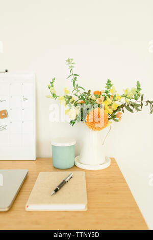 Hohe Betrachtungswinkel Tagebuch mit Stift durch Blumenvase auf hölzernen Schreibtisch gegen die Wand im Büro Stockfoto