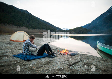 Freund, Freundin, beim sitzen auf der Picknickdecke am Seeufer von Lagerfeuer gegen den klaren Himmel Stockfoto