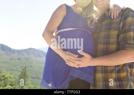 Man berühren die schwangere Frau Bauch, während sich gegen Berge während der sonnigen Tag Stockfoto