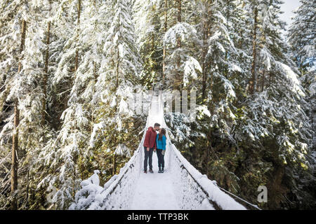 Paar küssen beim Stehen auf Steg inmitten von Bäumen im Wald am Lynn Canyon Park im Winter Stockfoto