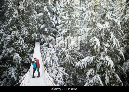 Volle Länge des Paar küssen beim Stehen auf Steg inmitten von Bäumen im Wald am Lynn Canyon Park im Winter Stockfoto