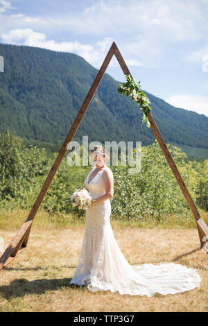 Braut holding Blumenstrauß beim Stehen auf Feld gegen Berg während der sonnigen Tag in Hochzeit Stockfoto