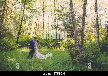 Frisch vermählte Paar küssen beim Stehen auf Wiese im Wald Stockfoto