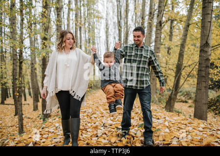 Verspielte Eltern schwingen Sohn, während seine Hände in Wald im Herbst Stockfoto