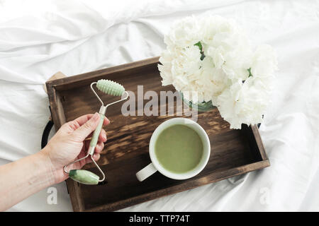 7/8 Hand der Frau mit Jade mit Matcha Tee und Blumen in Holz- Fach auf dem Bett Stockfoto