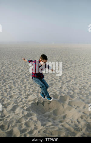 Verspielte junge Springen auf Sand am Strand gegen den klaren Himmel bei Sonnenuntergang Stockfoto