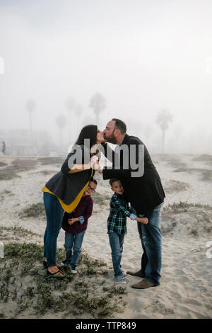 Seitenansicht der Eltern küssen beim Stehen mit den Söhnen am Strand während der nebligen Wetter Stockfoto