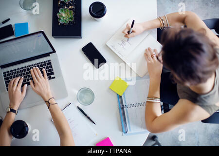 Ansicht von oben der Geschäftsfrau mit Laptop, während Kollege schreiben auf Buch im Büro Stockfoto