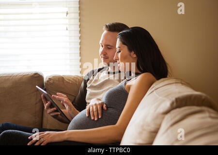 Mann mit Tablet Computer, während die schwangere Frau auf dem Sofa zu Hause sitzen Stockfoto