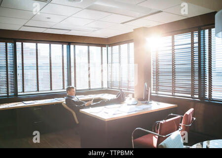 Geschäftsmann, der das Mobiltelefon benutzt, während er sich an sonnigen Tagen im Büro entspannt Stockfoto