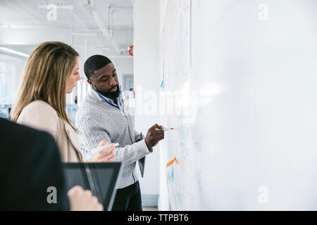 Geschäftsmann erklärt es einer Kollegin beim Schreiben auf Whiteboard im Büro Stockfoto