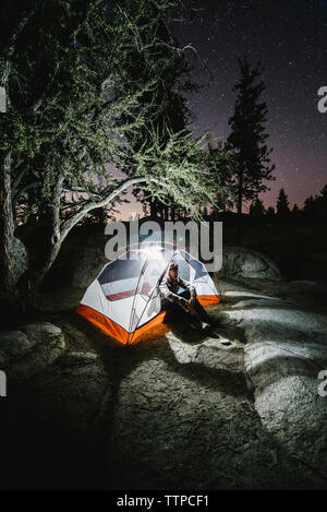 Wanderer sitzen in beleuchteten Zelt auf Rock von Bäumen in der Nacht Stockfoto