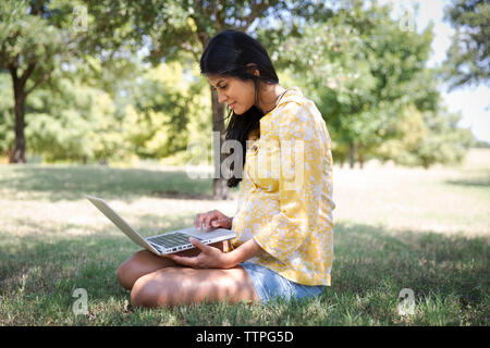 Lächelnde Frau mit Laptop, während auf dem Rasen im Park sitzen Stockfoto